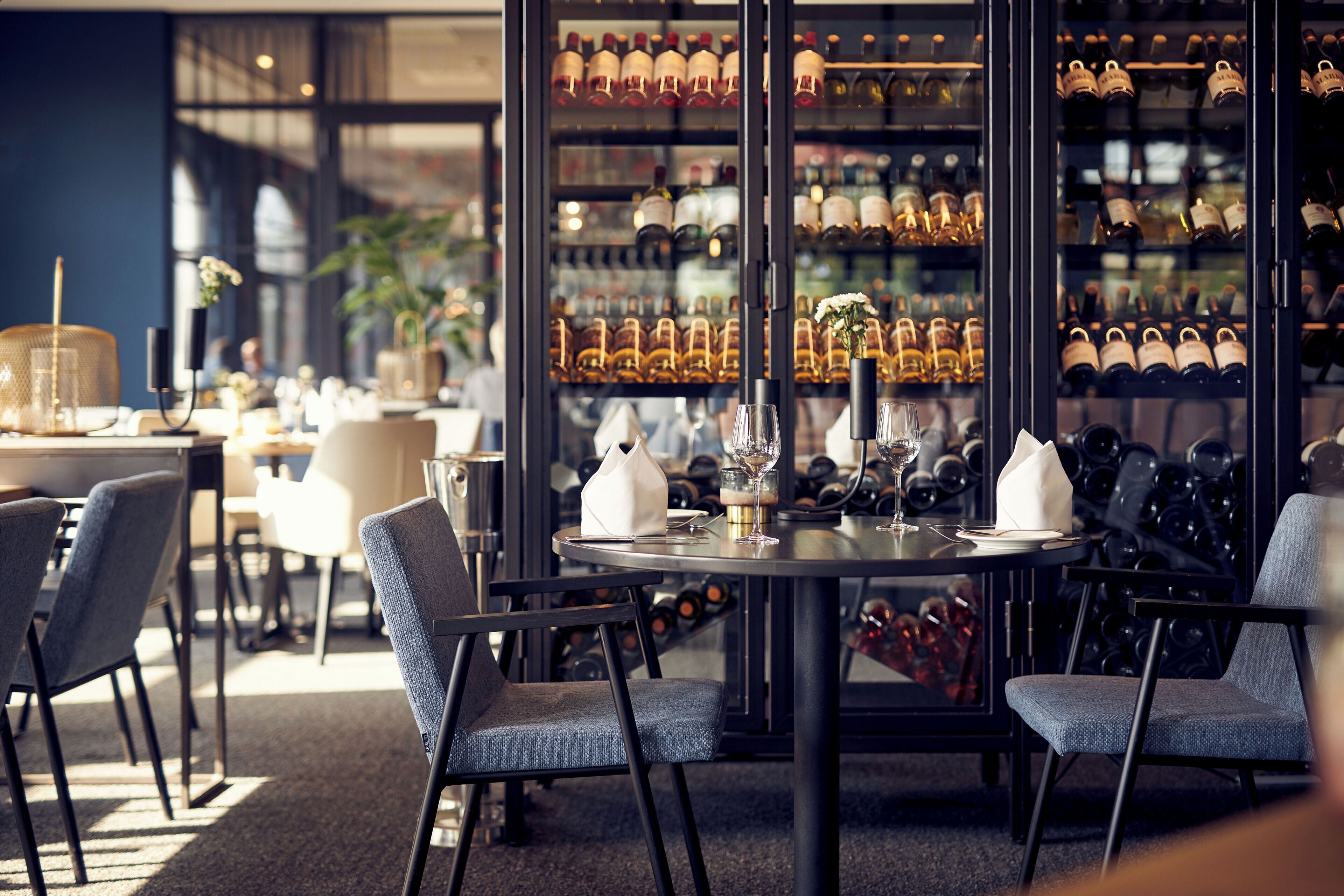 A restaurant interior with a round table, chairs, wine glasses, and a wine rack in the background.