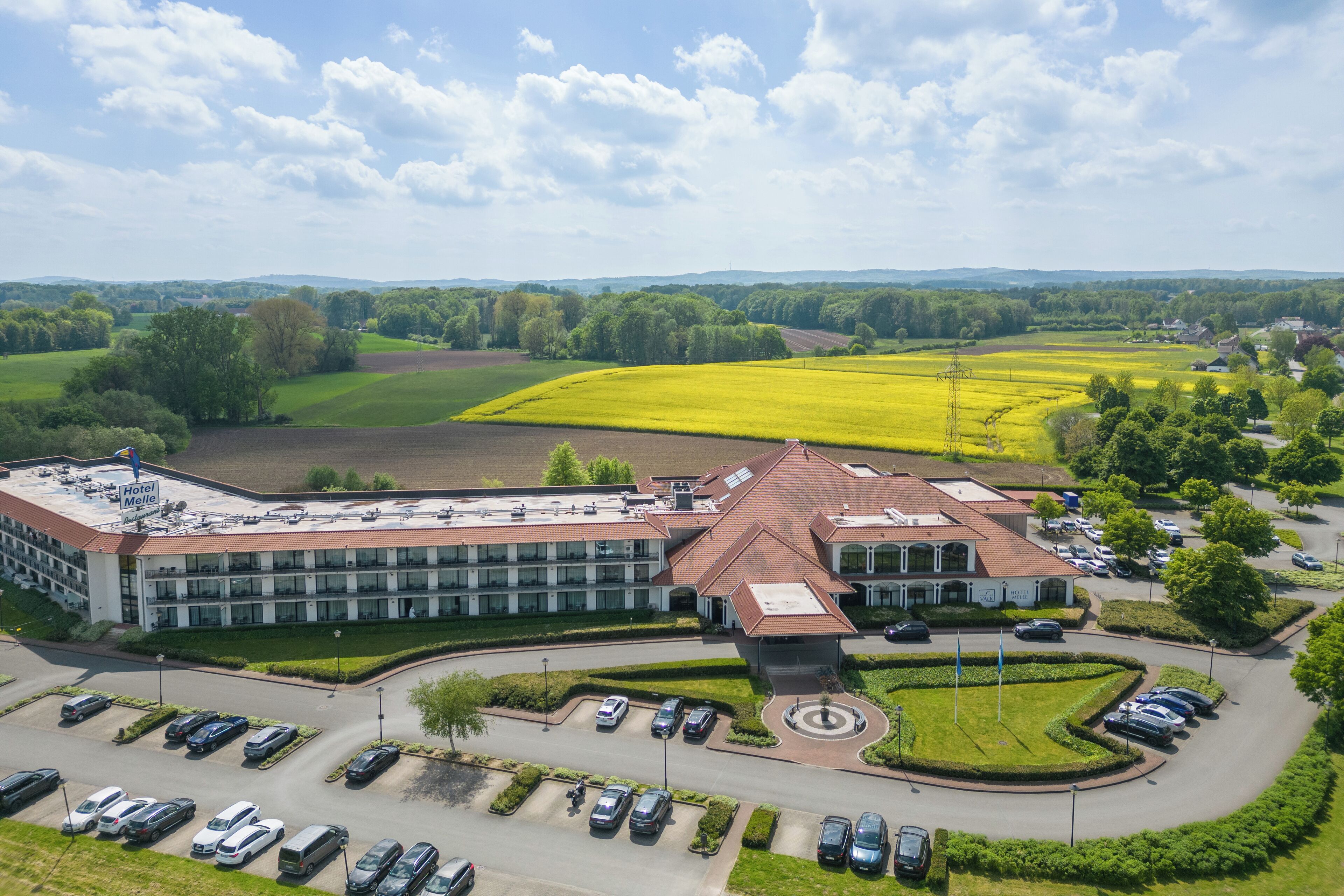 Un hotel grande con un patio central, rodeado de campos verdes y un estacionamiento con varios autos.