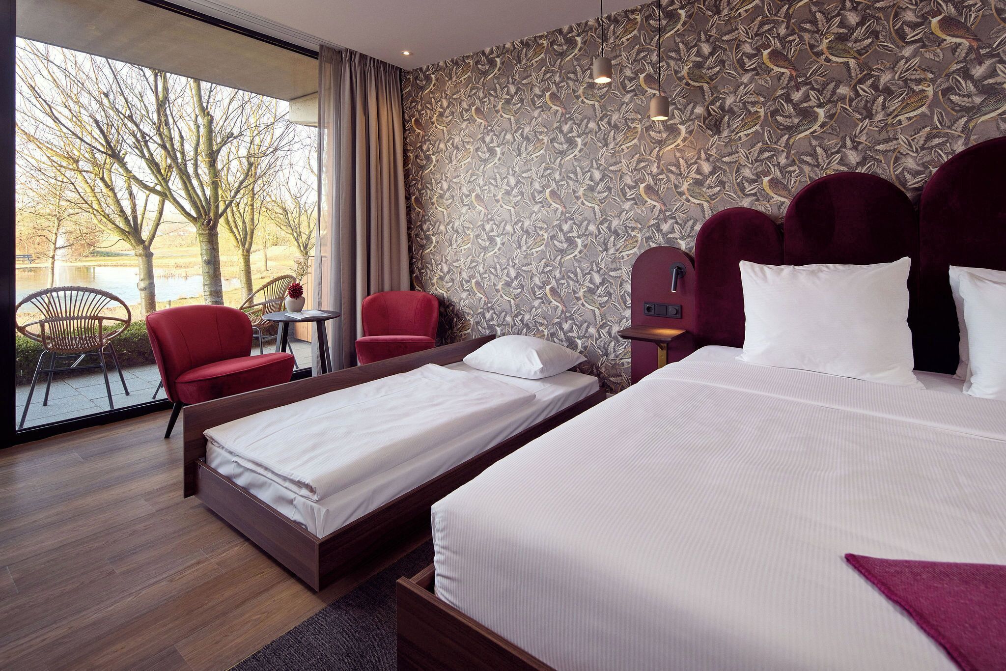 Habitación de hotel con dos camas, una silla roja, una mesita y vistas a árboles y agua.