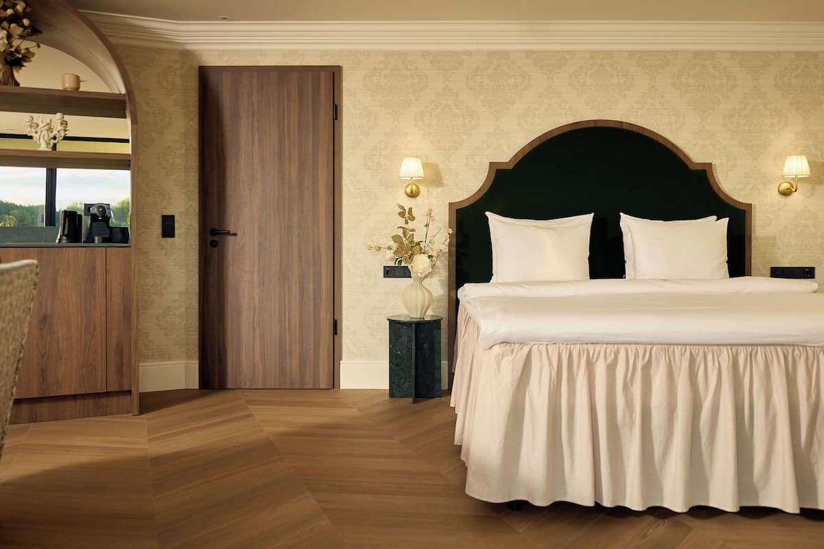 A bedroom with a large bed, wooden flooring, a dark headboard, two bedside lamps, and a small side table with a vase.