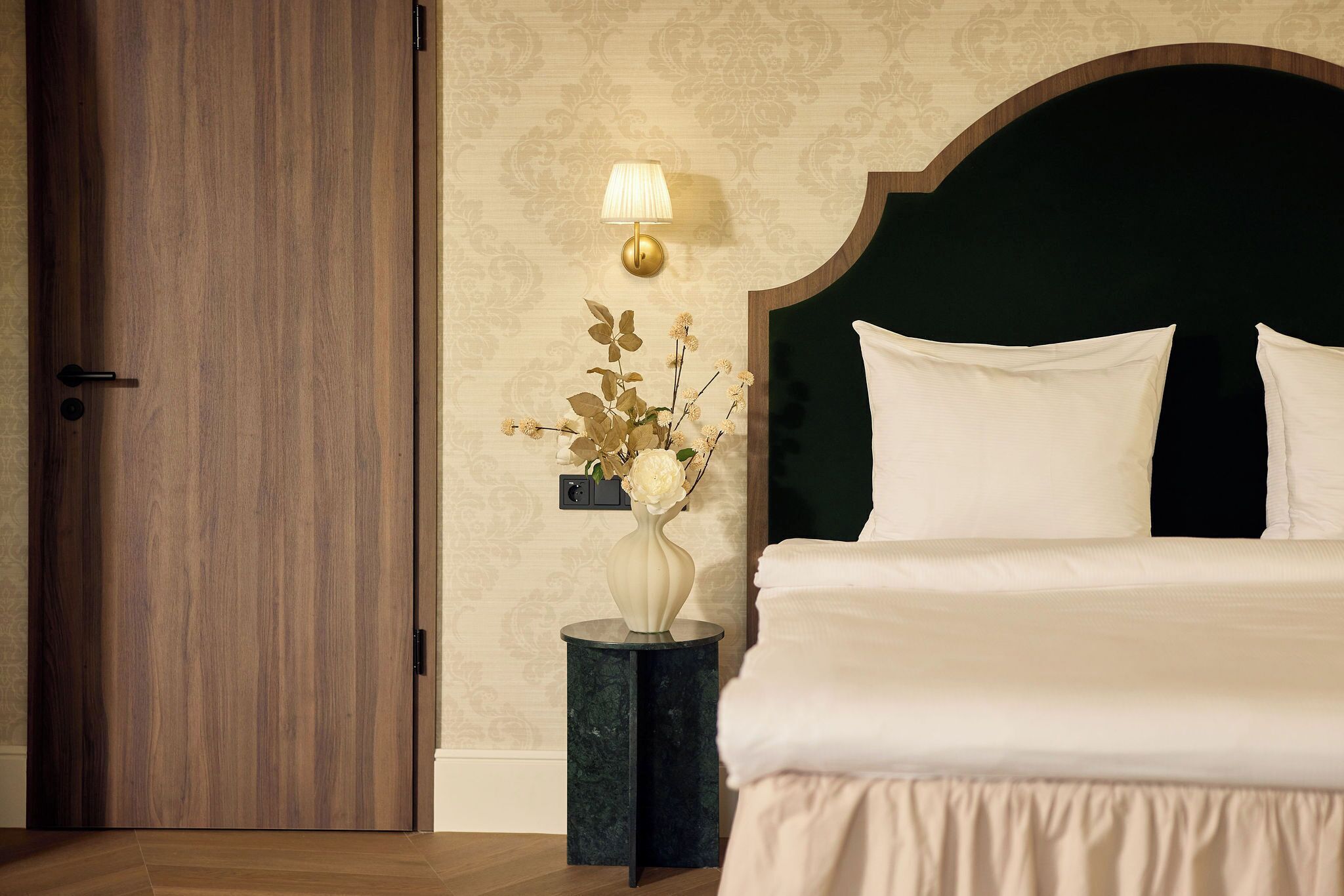A bedroom with a wooden door, a bedside table with a vase of flowers, a wall-mounted lamp, and a bed with white bedding.