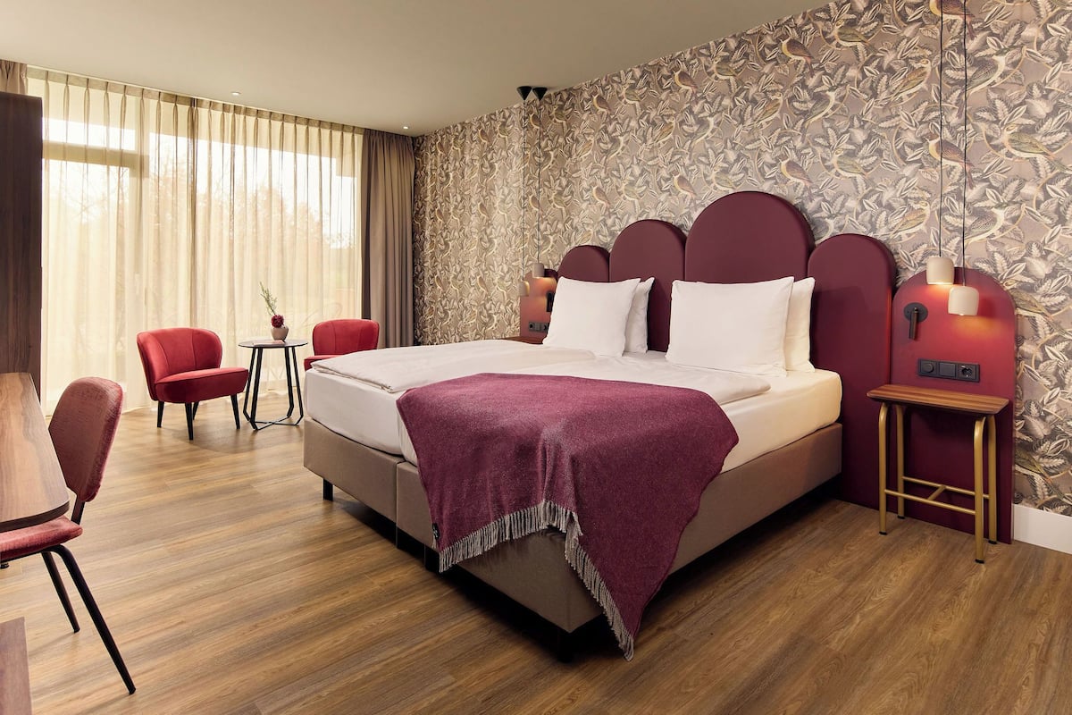 A hotel room with a large bed, red chairs, a small table, and a patterned wallpaper.