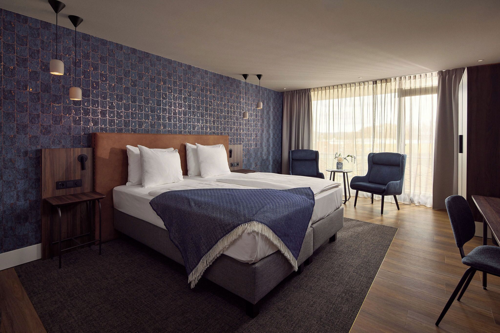 A modern hotel room with a large bed, a desk, two chairs, and a view of the outdoors.
