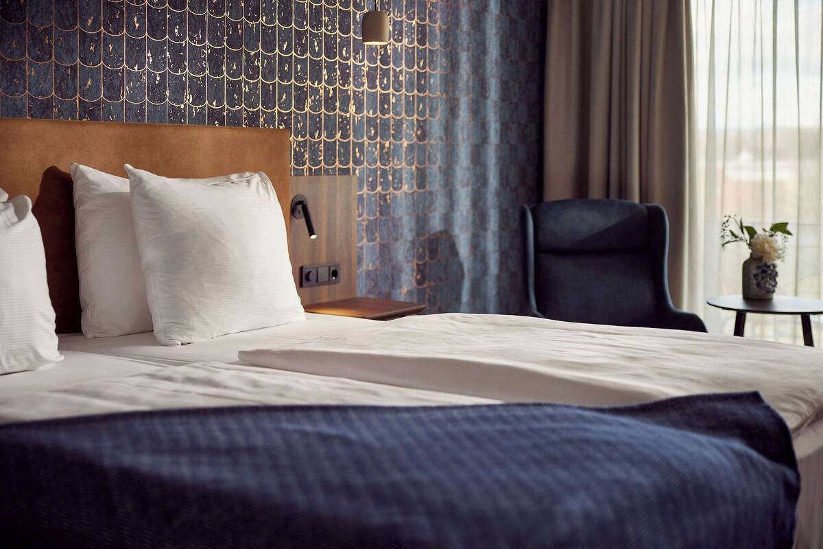 A hotel room with a bed, pillows, a headboard, a chair, a small table with a vase, and a wall with a unique pattern.