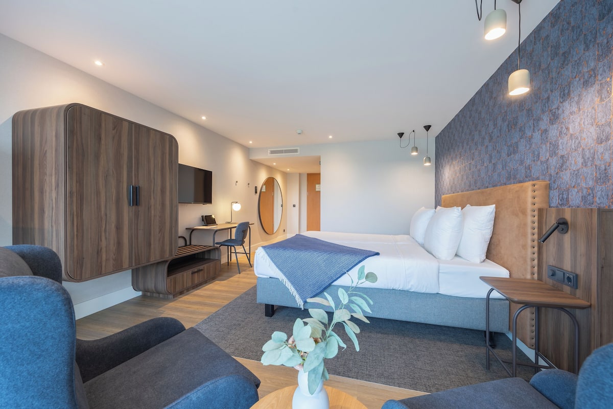 A modern hotel room with a large bed, a desk, a wardrobe, and a seating area with a plant.