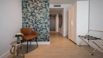 A modern hotel room with a wooden floor, a tropical leaf-patterned wall, a brown armchair, a small round table with a vase, and a foldable chair.