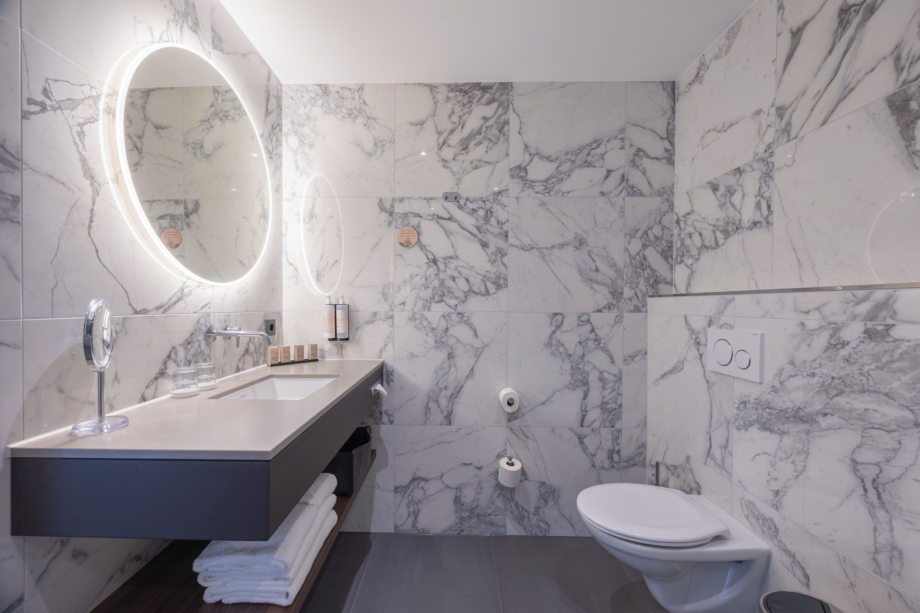 Une salle de bain moderne avec un pare-baignoire en marbre, un grand miroir doté d’un éclairage intégré, un lavabo blanc et des toilettes.