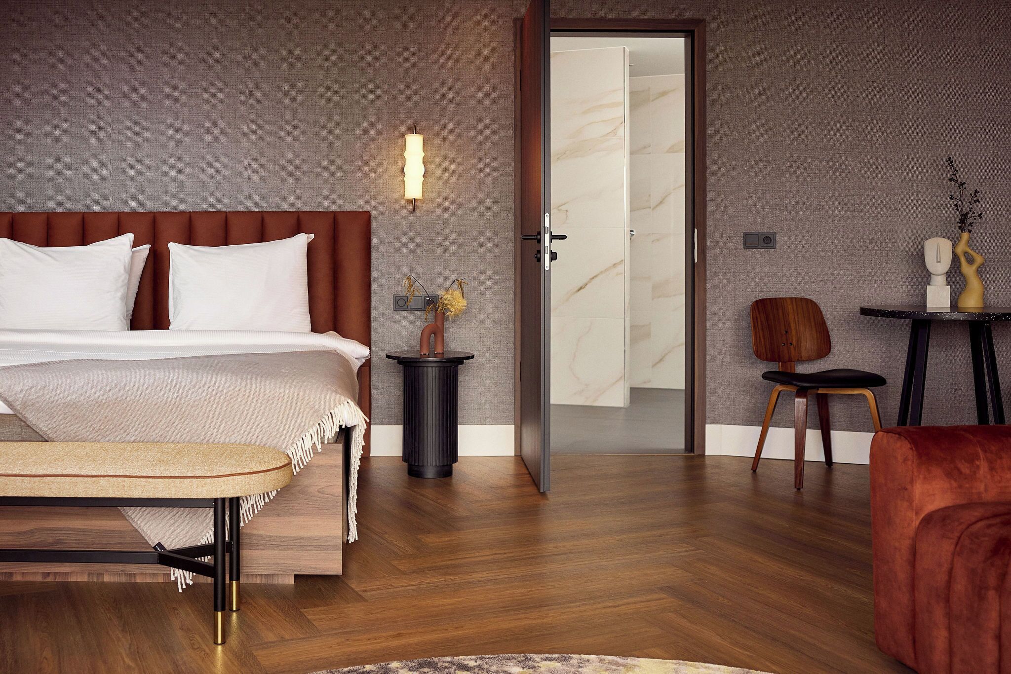 A modern hotel room with a bed, bedside table, chair, and a door leading to another room.