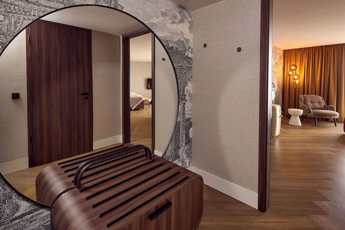 A modern bathroom with a wooden bench, a large mirror, and a view into a bedroom.