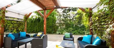 Un patio exterior con una pérgola, mobiliario de ratán negro y un toldo blanco.