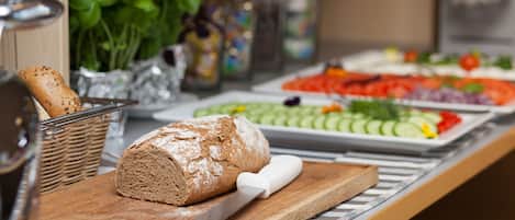 A buffet table with a loaf of bread, a knife, and various food items including sliced vegetables and a plate of meat.