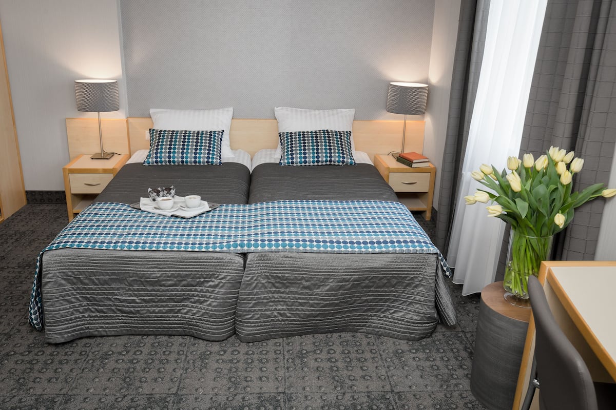 Una cama doble con colcha a cuadros, dos almohadas, una mesita de noche con lámpara, un florero con flores y una pequeña mesa con una silla.