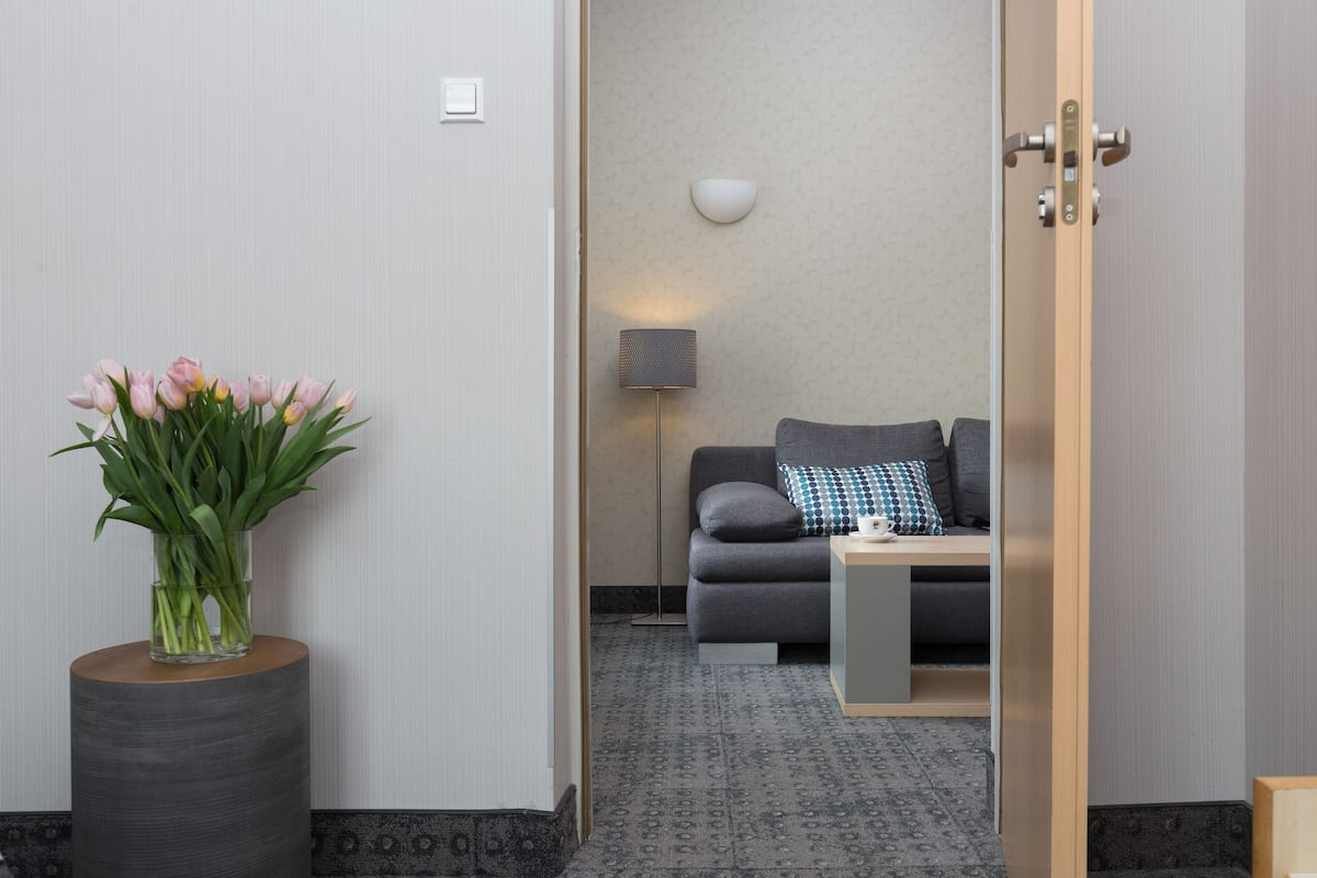 Una habitación de hotel moderna con un sofá gris, una mesita auxiliar pequeña y un jarrón con tulipanes rosados sobre una mesa redonda de madera.