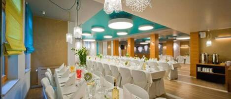 Un salón de banquetes con mesas redondas dispuestas para comer, sillas blancas y lámparas decorativas colgantes.