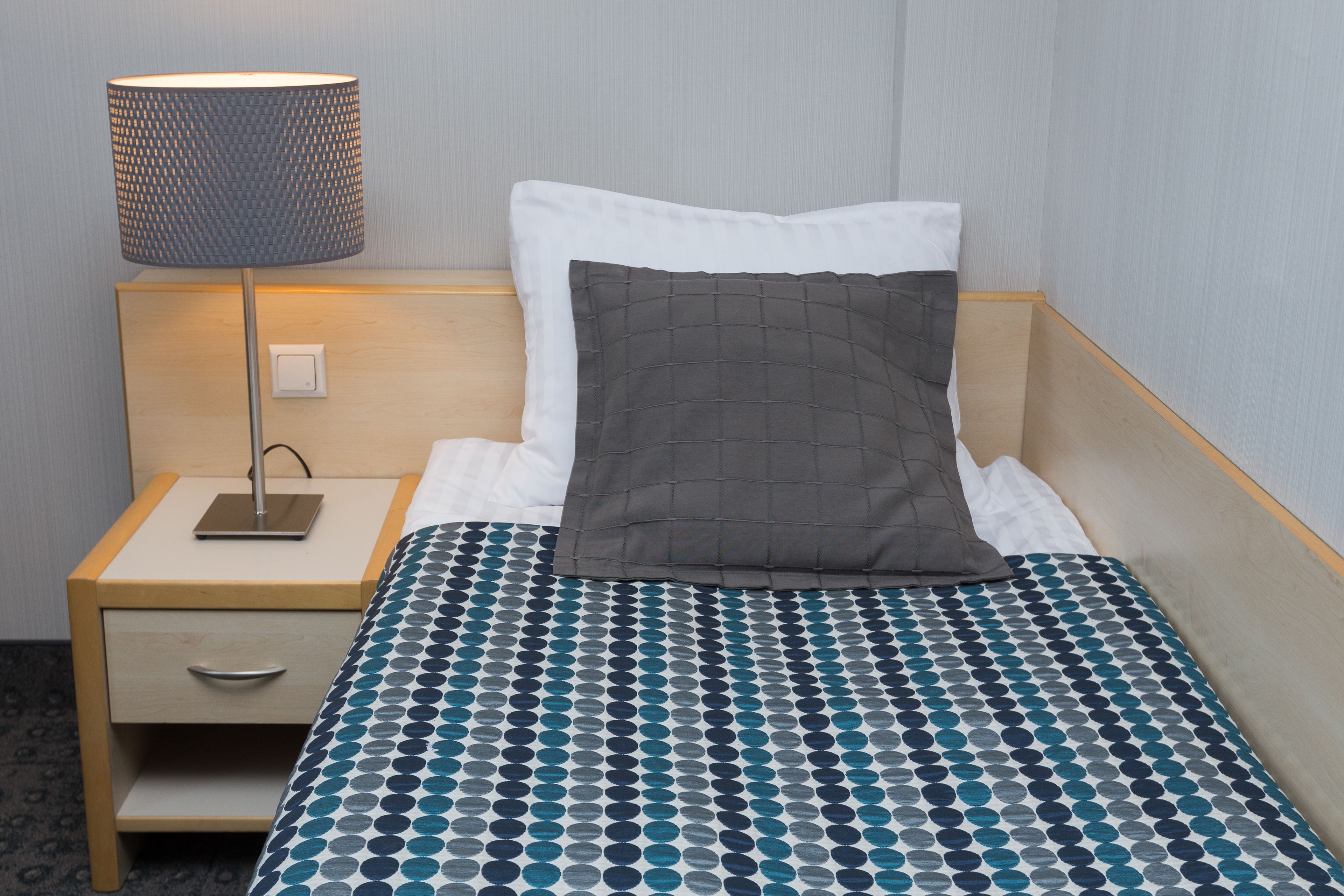 A neatly made bed with a blue and white polka dot bedspread, a gray quilted pillow, and a bedside lamp on a wooden nightstand with a drawer.