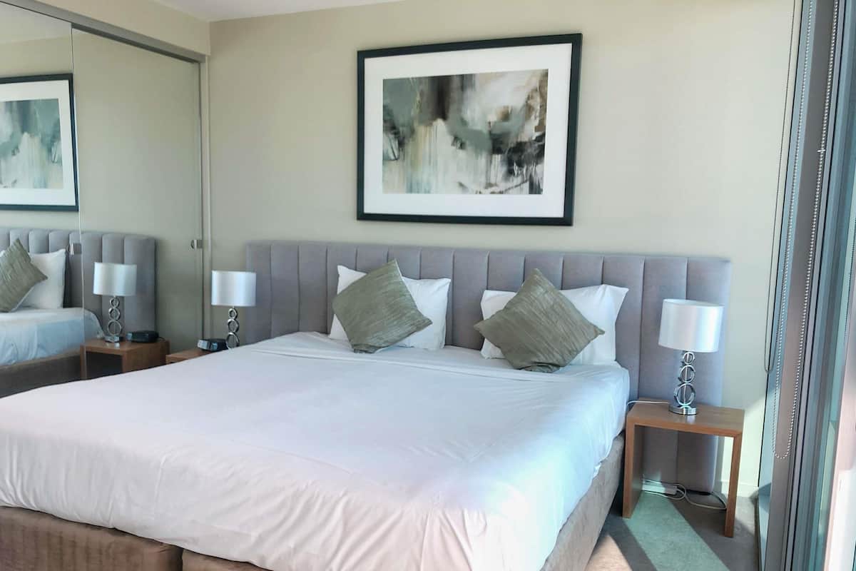 Ein Hotelzimmer mit einem großen Bett, zwei Nachttischen mit Lampen, einem gerahmten abstrakten Gemälde an der Wand und einem großen Spiegel über dem Bett.
