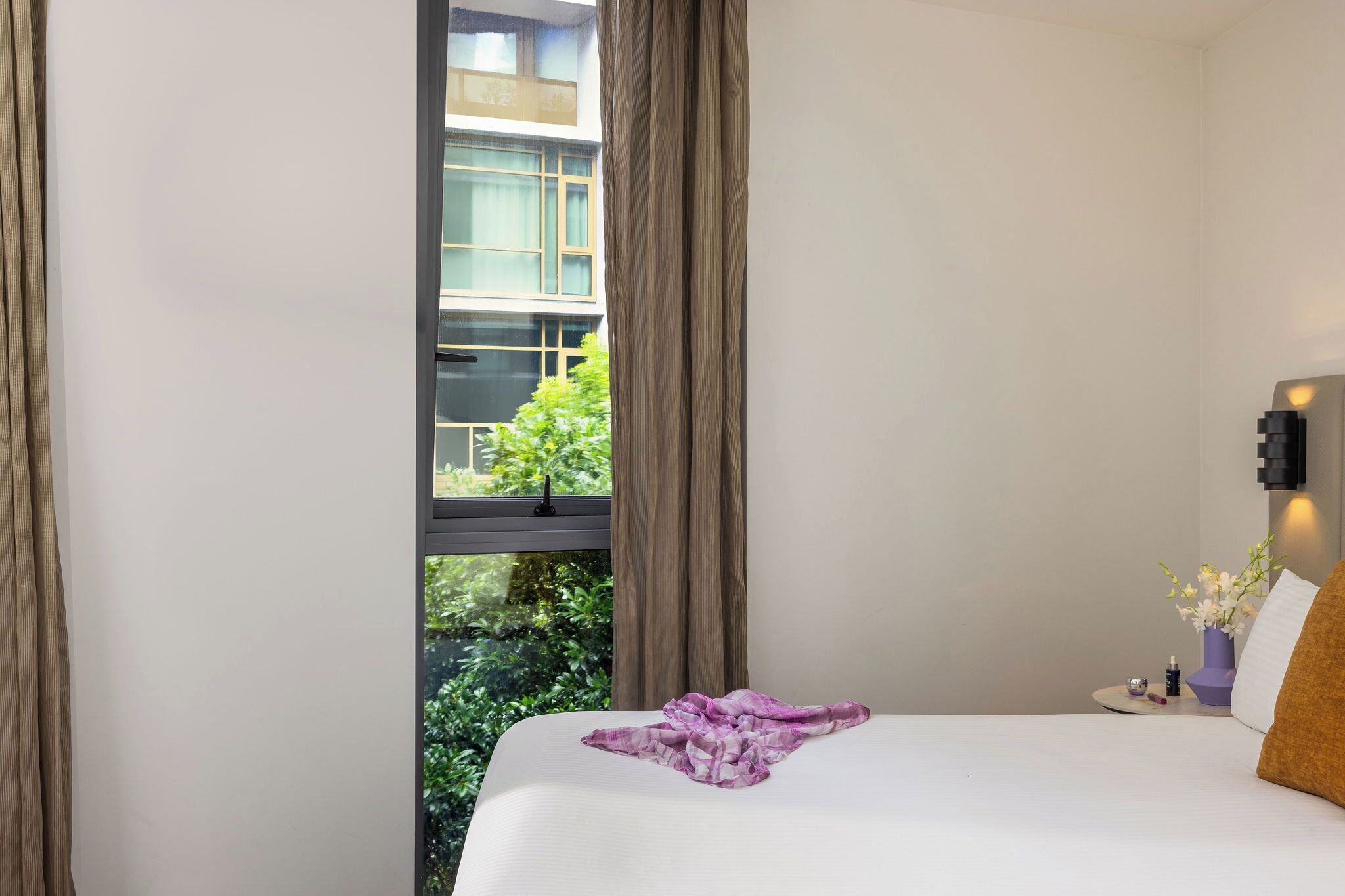 A hotel room with a bed, a nightstand, a window with curtains, and a view of greenery outside.