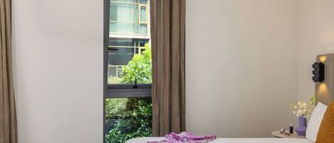 A hotel room with a bed, a nightstand, a window with curtains, and a view of greenery outside.