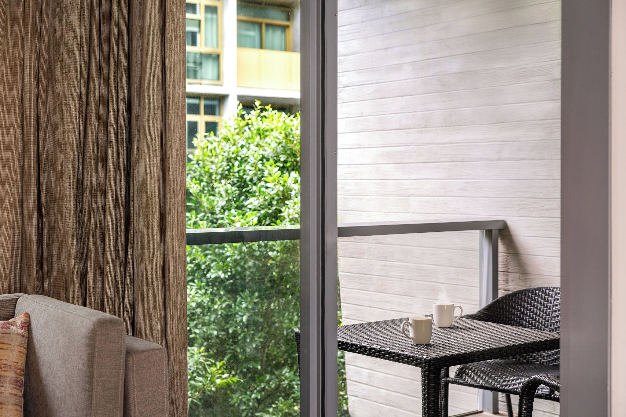A balcony with a wicker table and two white cups, a grey sofa, and a view of greenery and buildings.