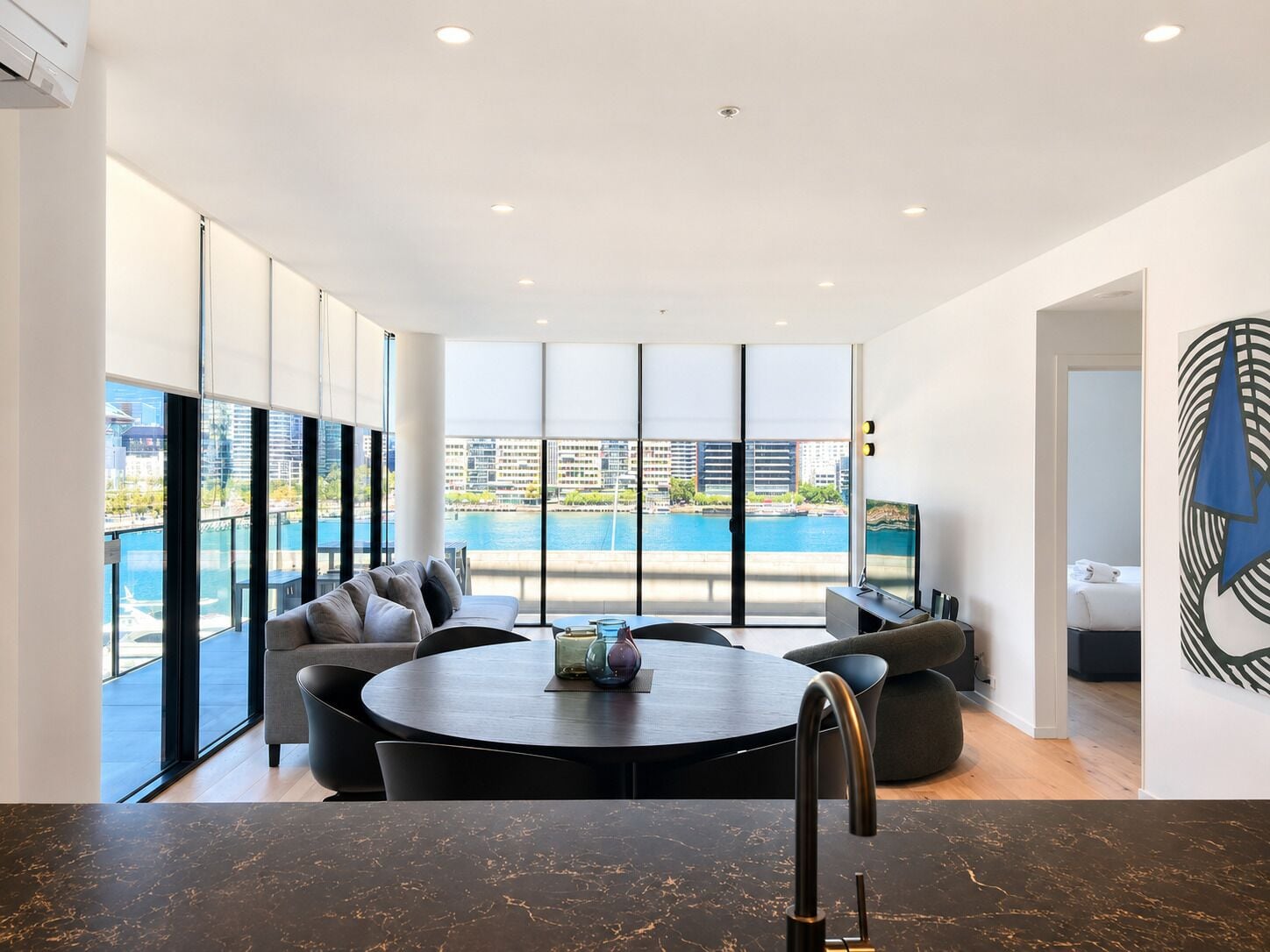 Una sala de estar moderna con una mesa redonda, asientos y un amplio ventanal que ofrece vistas a un cuerpo de agua y edificios.