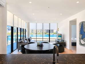 A modern living room with a round table, seating, and a large window offering a view of a body of water and buildings. - The Sebel Residences Melbourne Docklands Serviced Apartments (Docklands)