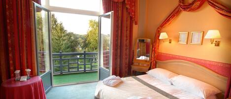 A hotel room with a large bed, a small table with a red tablecloth, two wall-mounted lamps, and a view of greenery through the window.
