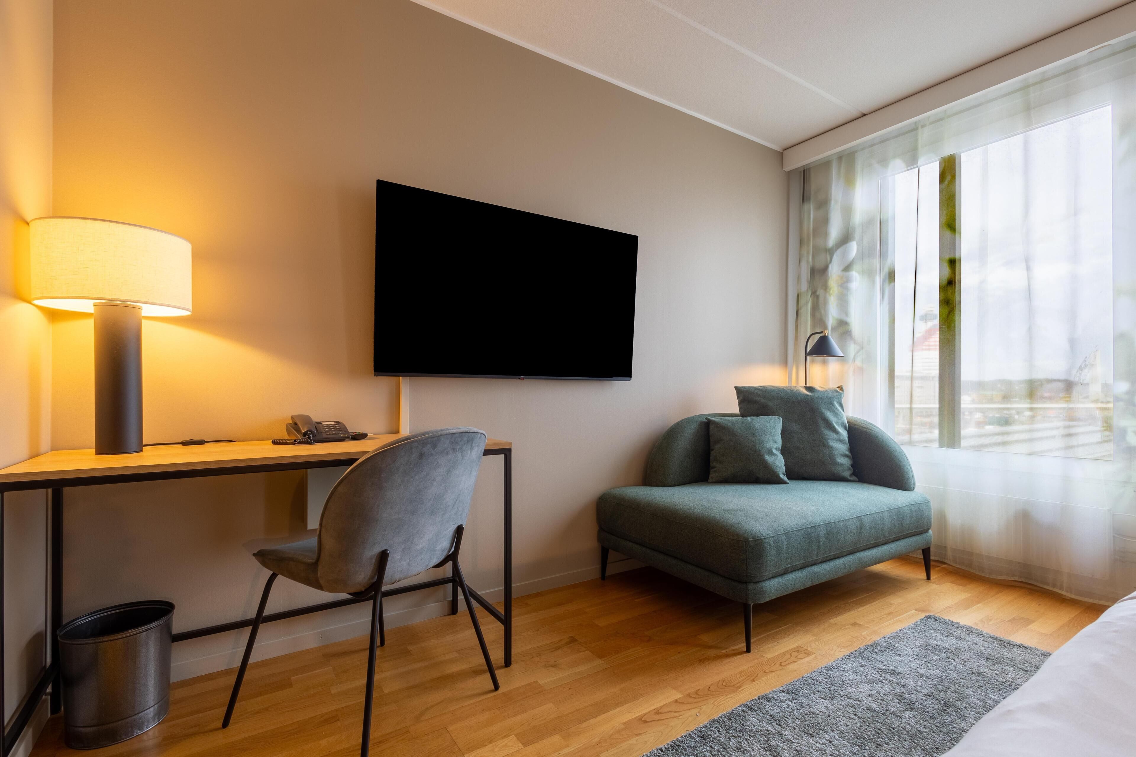 Quarto de hotel moderno com escrivaninha, cadeira, sofá e uma TV de tela plana na parede.