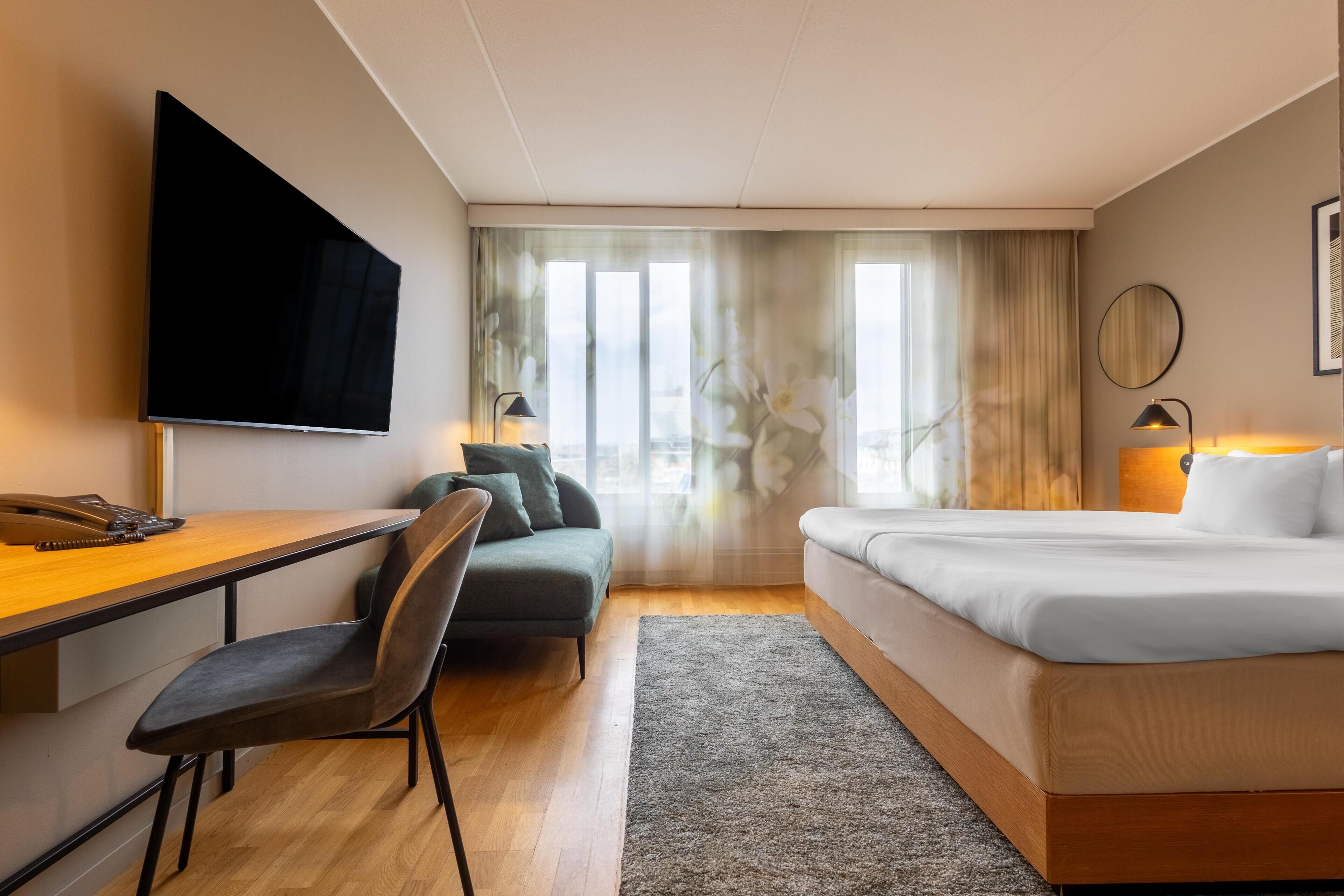 A hotel room with a flat-screen TV, a desk with a telephone, a sofa, a bed, and a window with curtains.
