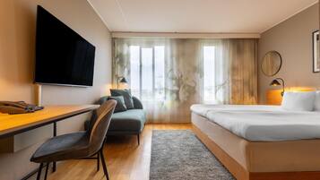 A hotel room with a flat-screen TV, a desk with a telephone, a sofa, a bed, and a window with curtains.