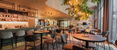 Zona de bar moderna con mesas y sillas redondas, barra con varias botellas y copas, y planta colgante con luces.