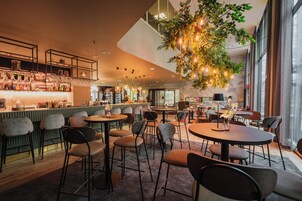 Zona de bar moderna con mesas y sillas redondas, barra con varias botellas y copas, y planta colgante con luces.
