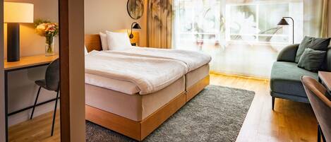 Habitación de hotel moderna con una cama grande, un escritorio con lámpara, una silla y vistas al exterior a través de amplios ventanales.