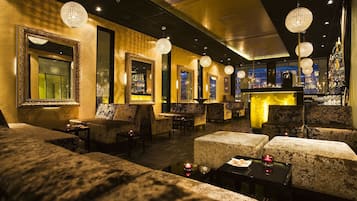 Ett svagt upplyst loungeområde med bekväma sittplatser, kristallkronor och en bar i bakgrunden.