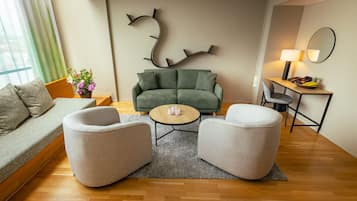 Un salon moderne avec un canapé vert, deux fauteuils, une table basse et une table d’appoint sur laquelle se trouvent une lampe et un panier de fruits.