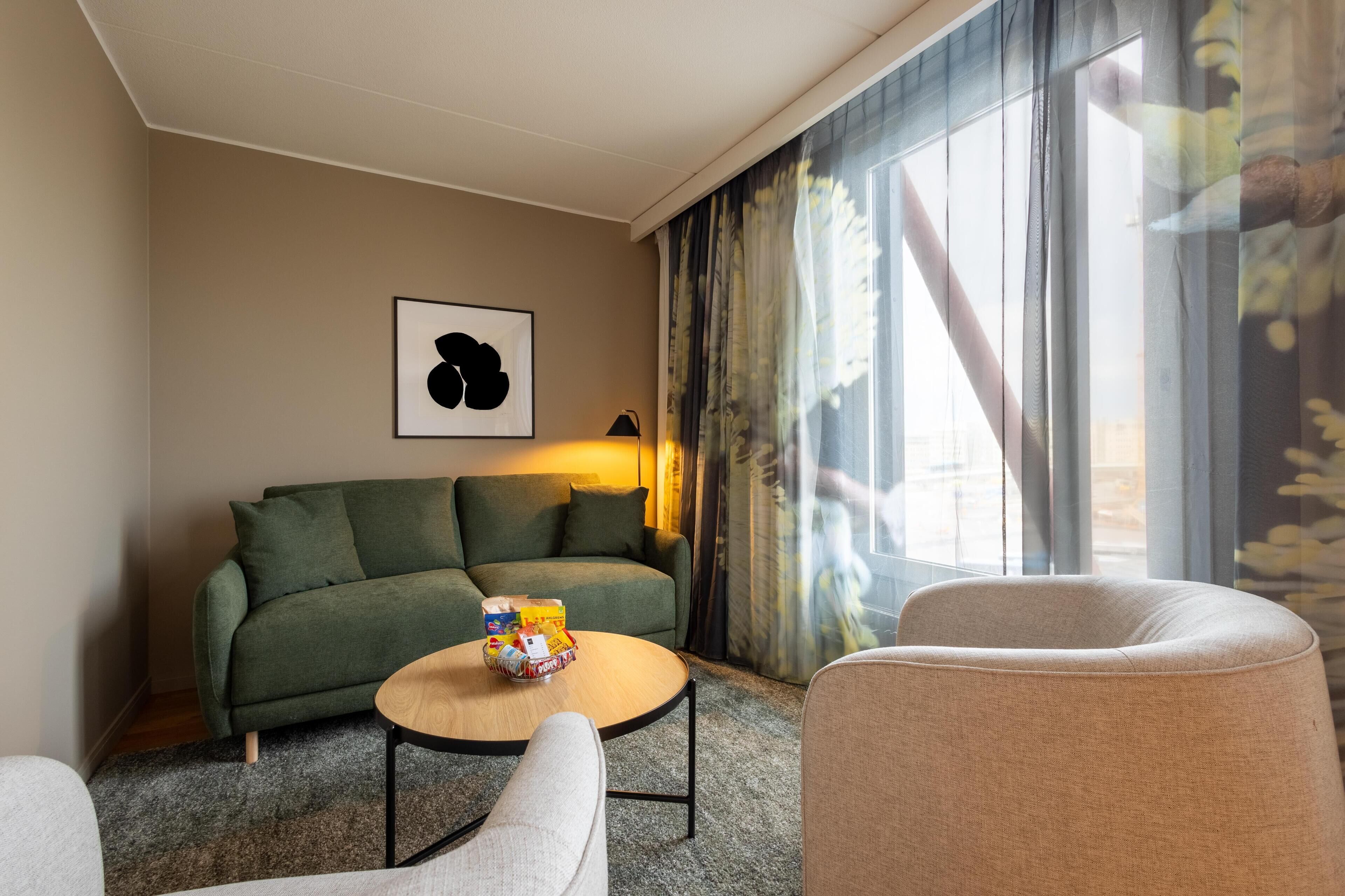 Una habitación de hotel moderna con un sofá verde, una mesa de centro redonda con aperitivos y un ventanal con cortinas.