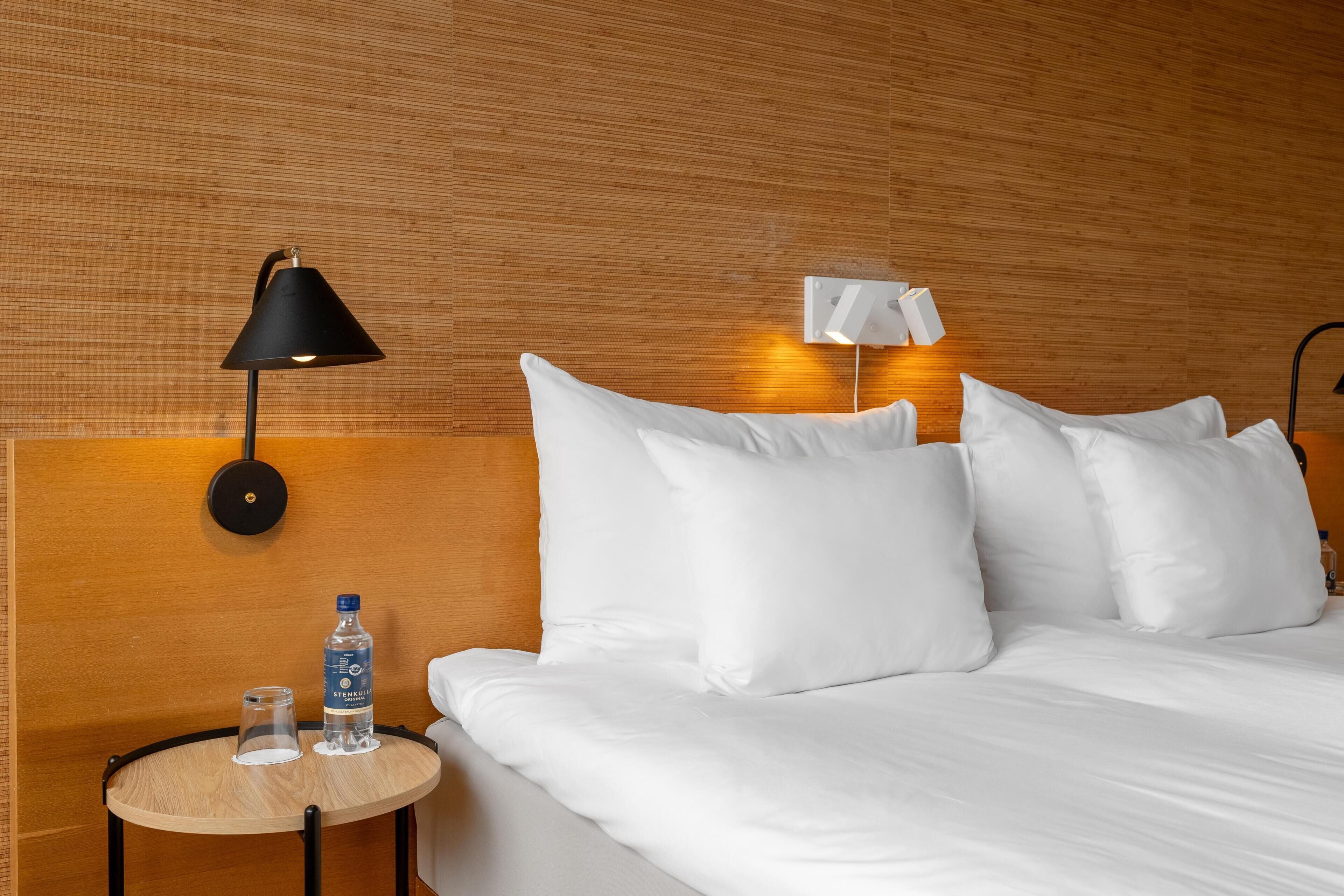 A hotel room with a bed, a round table with a bottle and glasses, a wall-mounted lamp, and a headboard with wall lights.