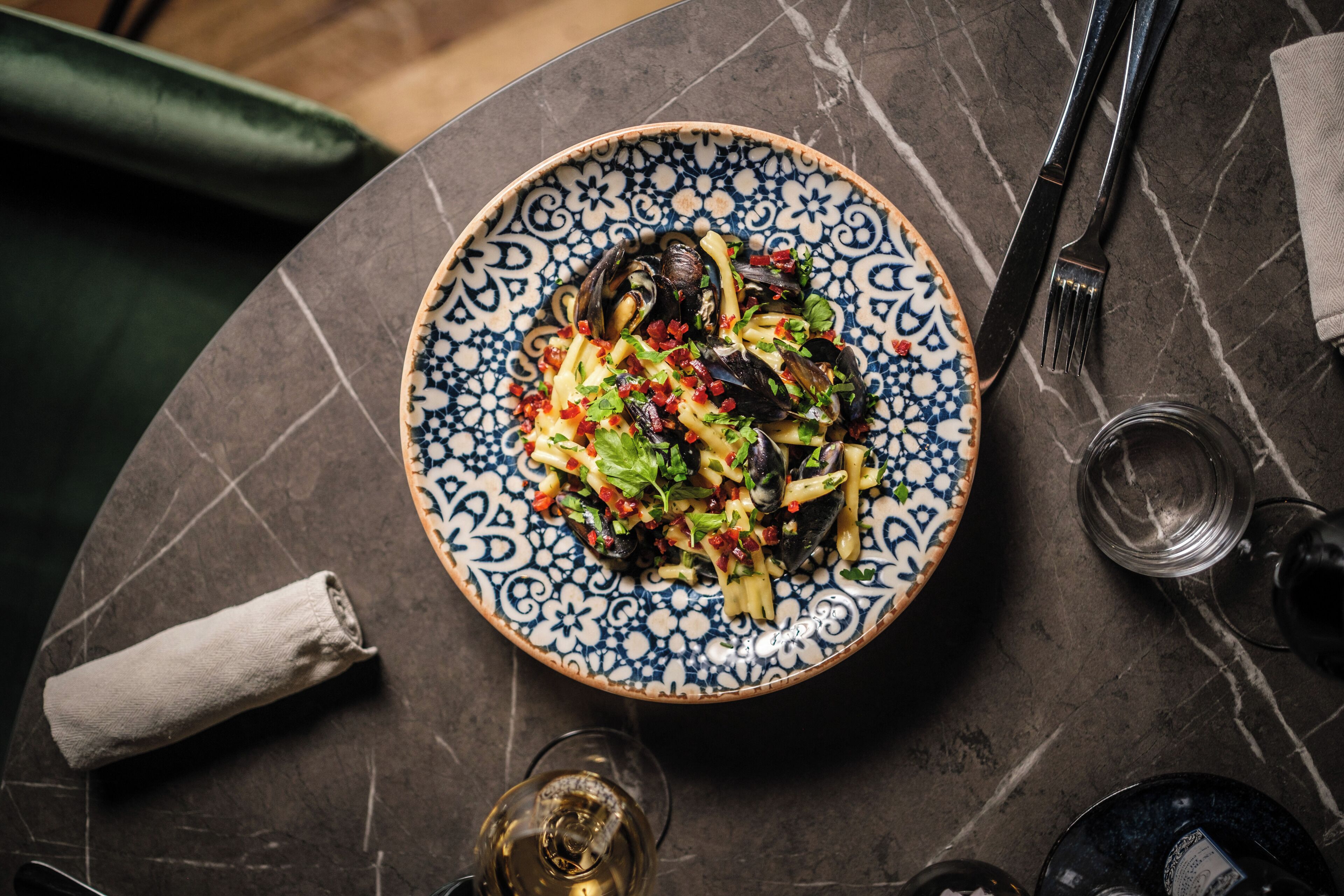 Una mesa con un plato de pasta, un tenedor, un cuchillo, una servilleta y una copa de vino.