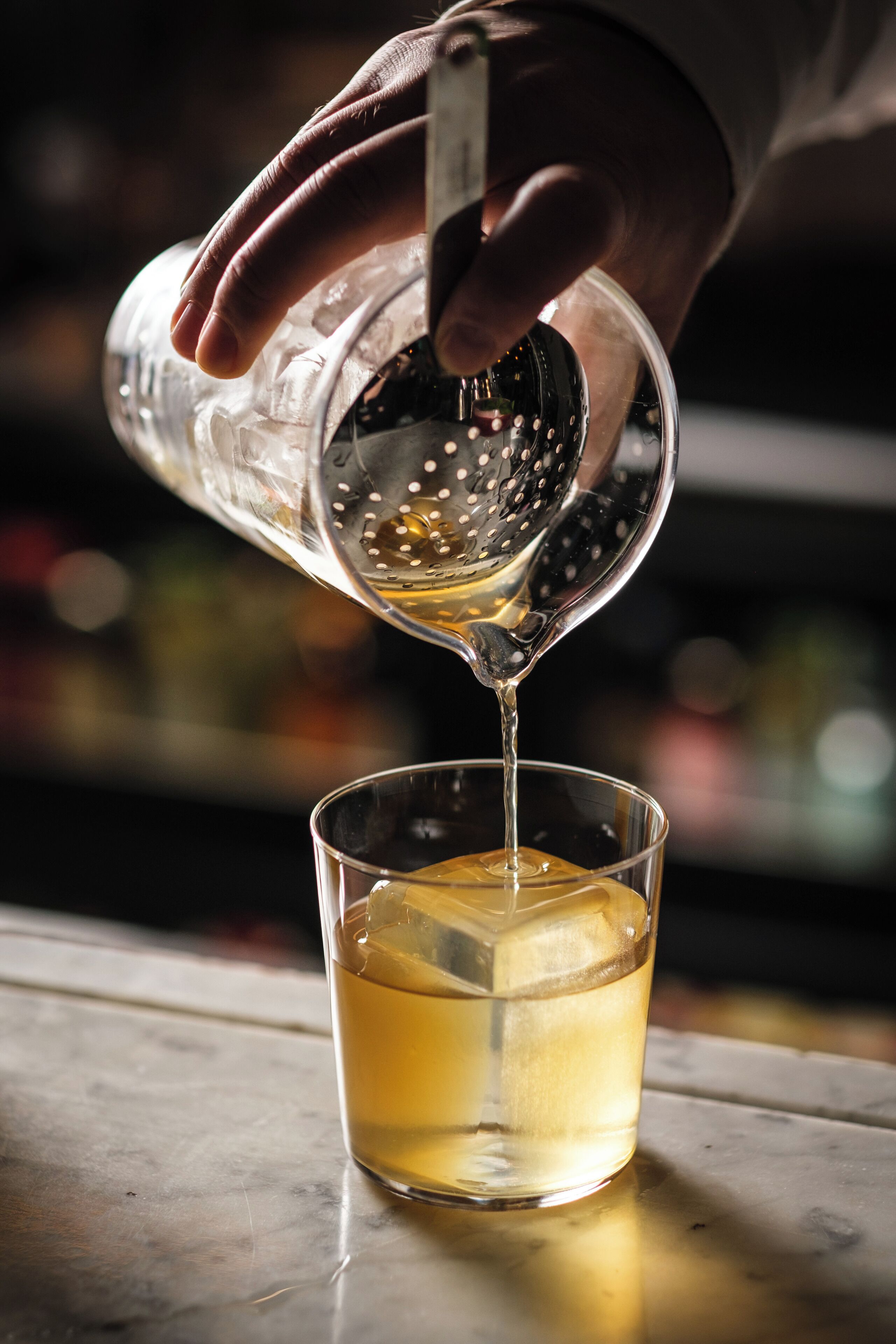 A hand pouring a clear liquid into a glass with an ice cube.