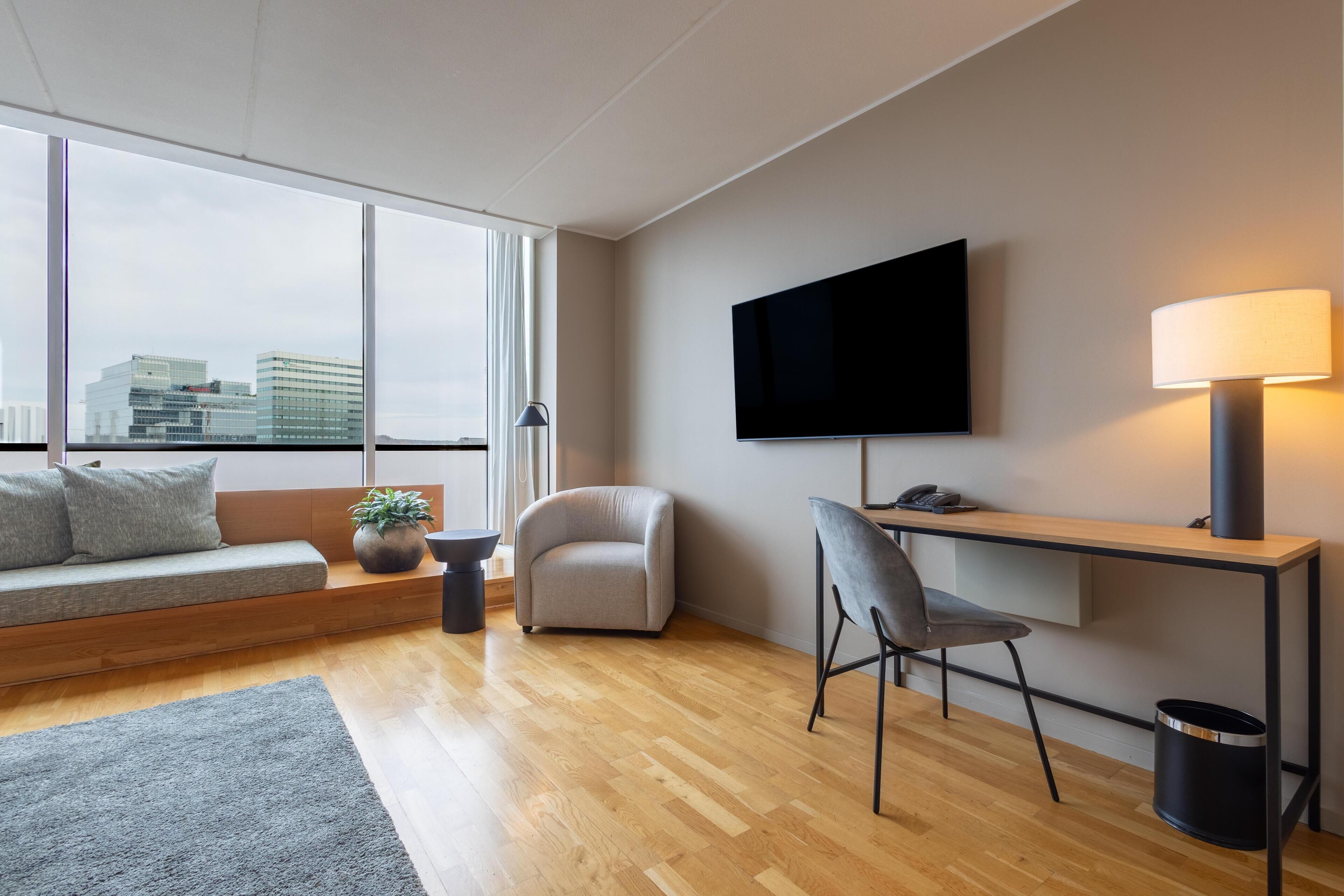 A modern living room with a sofa, armchair, and a desk with a chair. A flat-screen TV is mounted on the wall, and a lamp is on the desk.