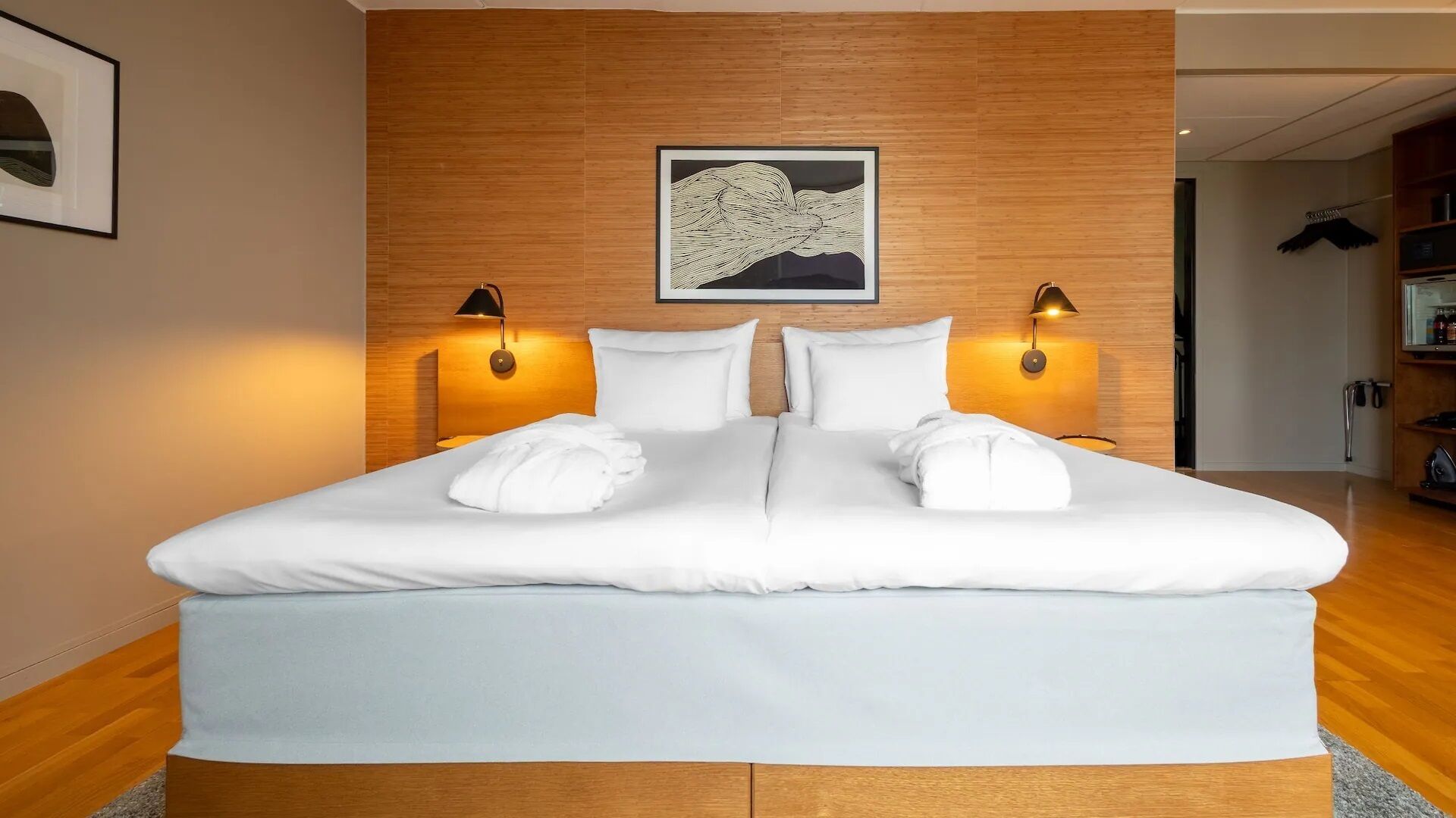 A double bed with white linens, two bedside lamps, a framed artwork above the bed, and a wooden headboard.