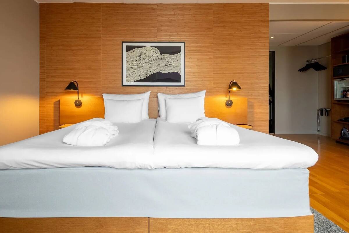 A double bed with white linens, two bedside lamps, a framed artwork above the bed, and a wooden headboard.
