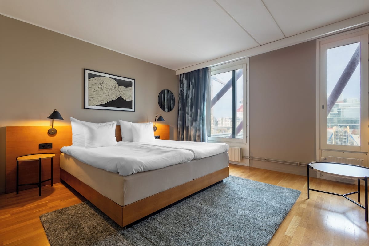 Ett modernt sovrum med en stor säng, sänglampor, ett litet bord och utsikt över staden genom stora fönster.
