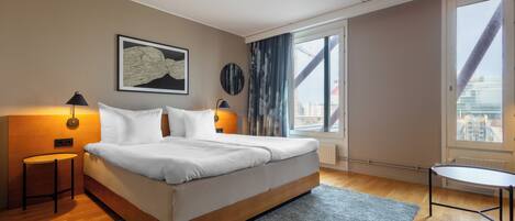 A modern bedroom with a large bed, bedside lamps, a small table, and a view of the city through large windows.