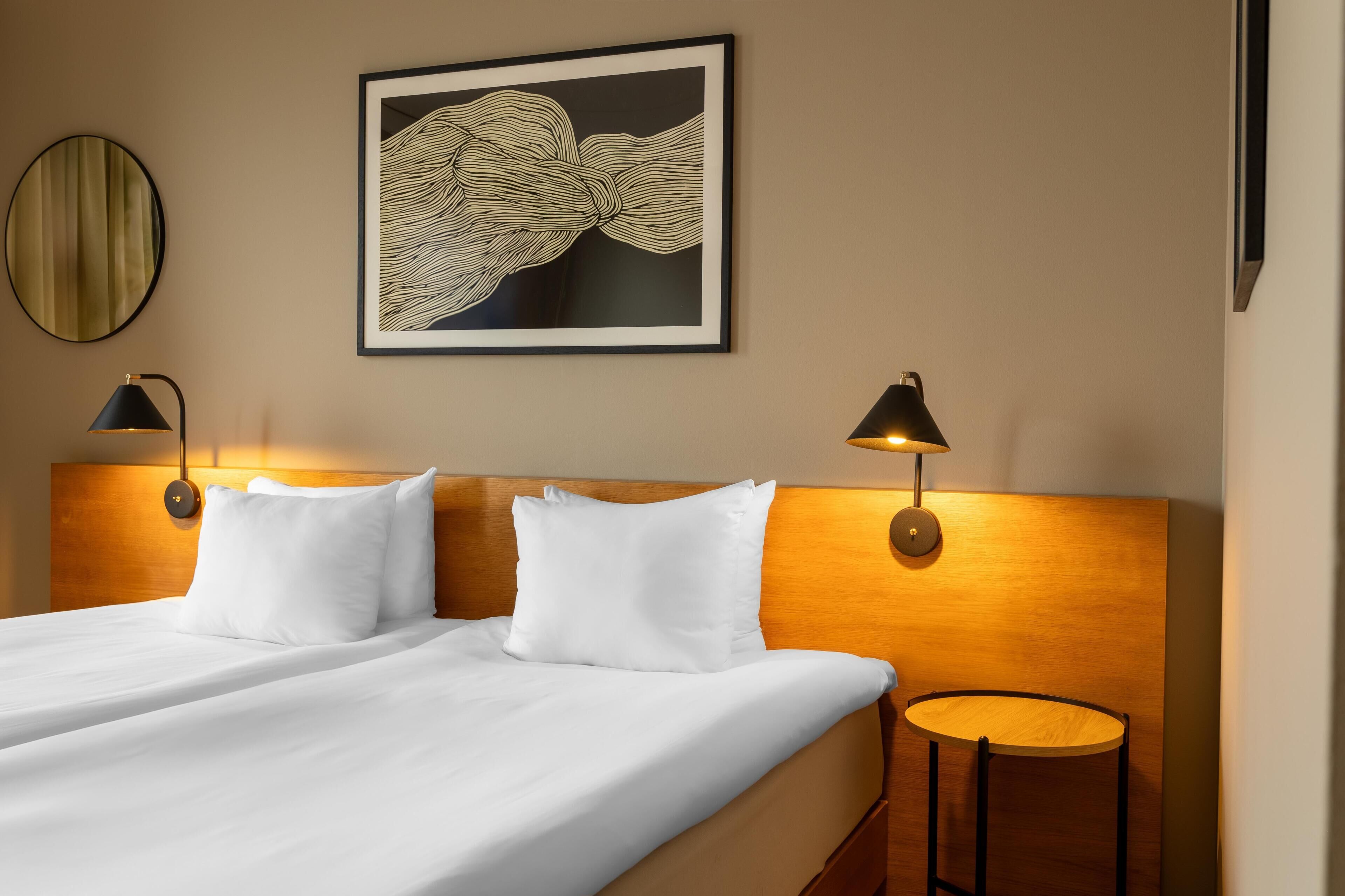 A hotel room with a neatly made bed, two bedside lamps, a round table, and a framed artwork on the wall.