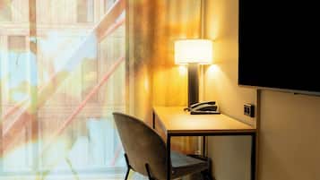 A hotel room with a desk, chair, lamp, and a large window with a view of a building.