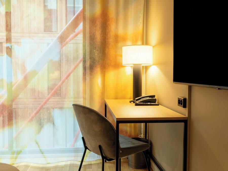 Quarto de hotel com escrivaninha, cadeira, abajur e uma janela grande com vista para um edifício.