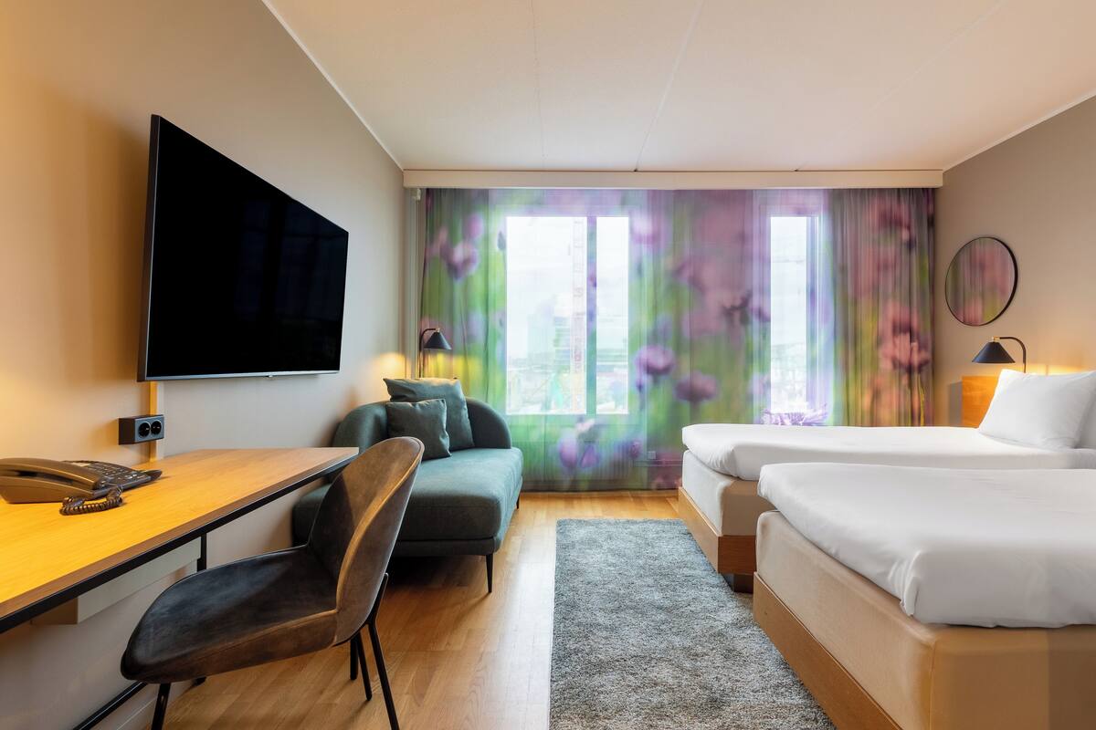 Ett hotellrum med ett skrivbord, en stol, en soffa, två sängar, en TV och ett f önster med gardiner.
