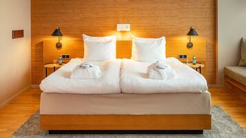 Een moderne hotelkamer met een houten hoofdeinde, een groot bed met witte lakens, twee nachtkastjes met lampen, en een klein tafeltje met een fles en glas.