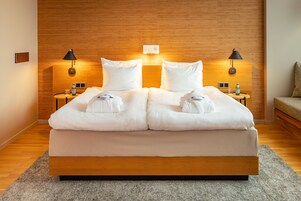 Una habitación de hotel moderna con cabecera de madera, una cama grande con ropa blanca, dos mesitas de noche con lámparas, y una pequeña mesa con una botella y un vaso.