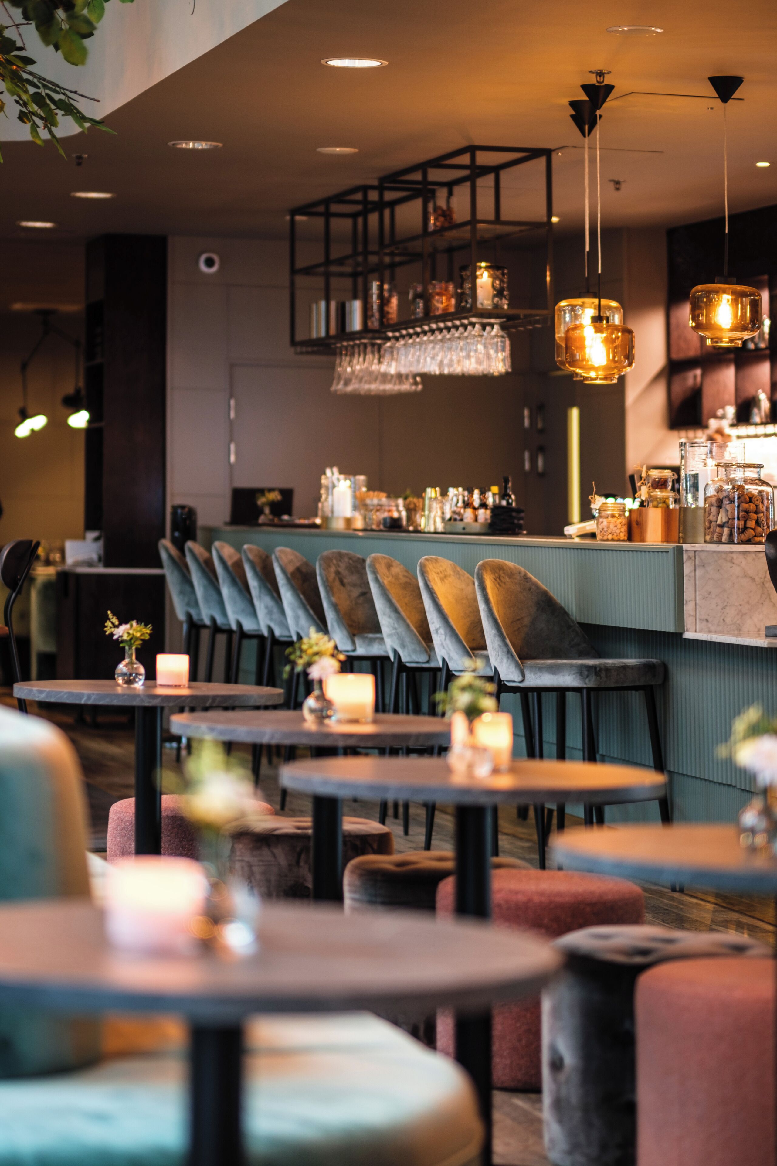 Interior de un restaurante con varias mesas y sillas, una zona de bar con estantes llenos de botellas y lámparas colgantes.